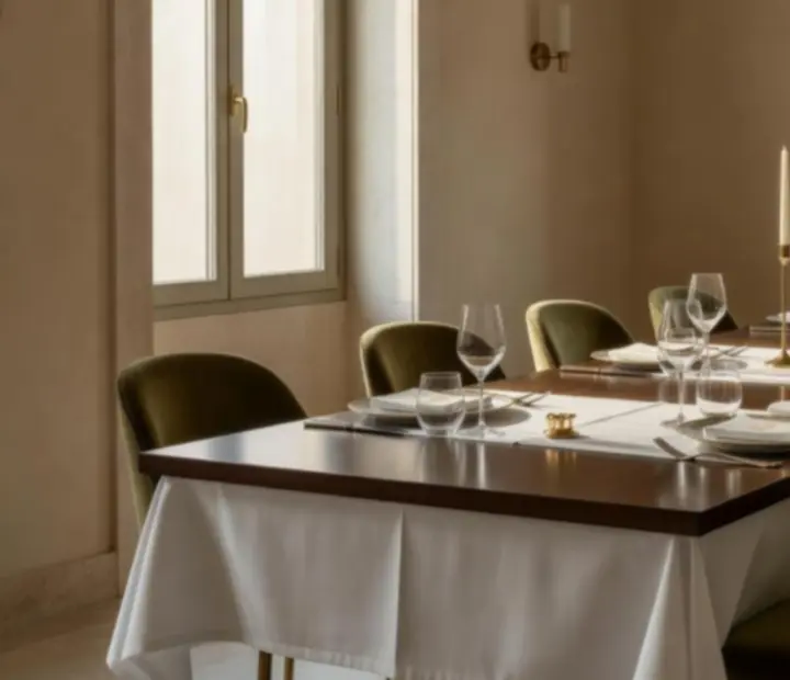 Mesa preparada con mantel blanco, copas de vino y detalles de cartas junto a ventanales con luz mediterránea.